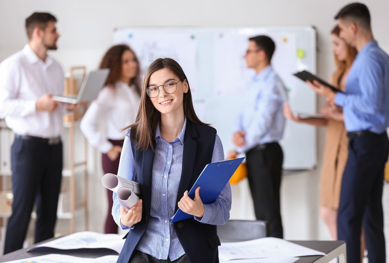 A young woman standing in an office facing the camera and smiling while holding a clipboard and some documents. Behind her five men and women are involved in a business discussion.