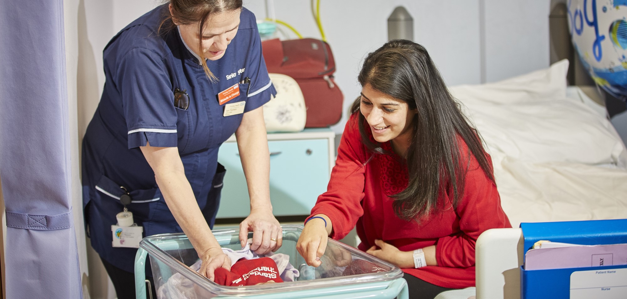 A smiling mother and a midwife looking at a newborn baby in its bedside cot